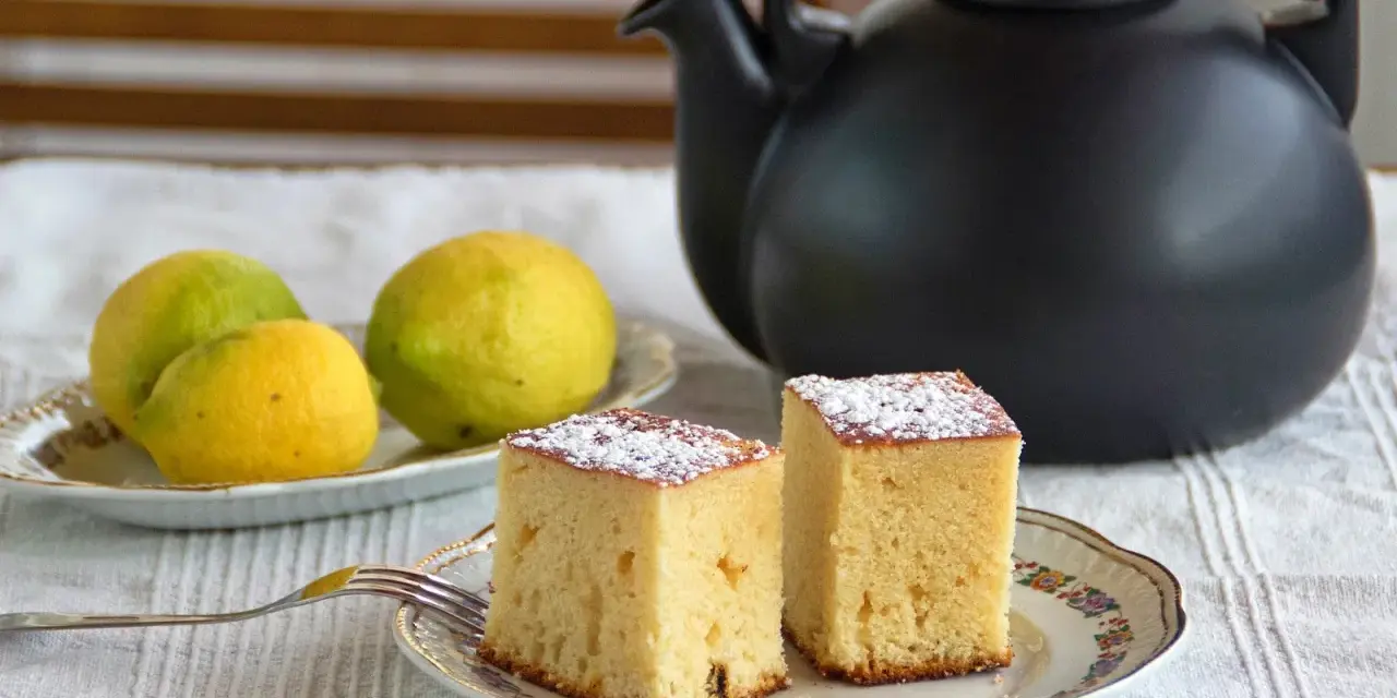 Dos porciones de bizcocho de limón espolvoreadas con azúcar glas, junto a limones frescos y una tetera negra.