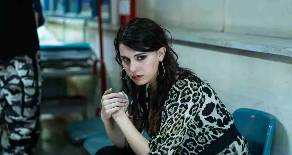 Carmela, con sguardo intenso, attende il suo destino fuori dal carcere. Indossa una camicia leopardata.
