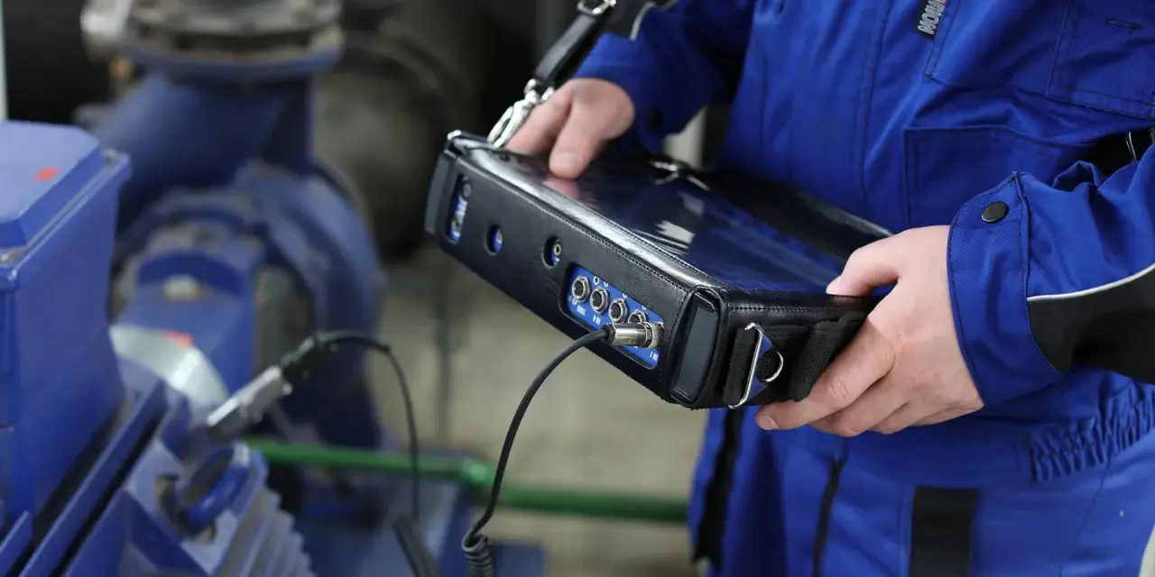 Technik w niebieskim kombinezonie wykonuje pomiary drgań maszyny przemysłowej za pomocą przenośnego urządzenia.
