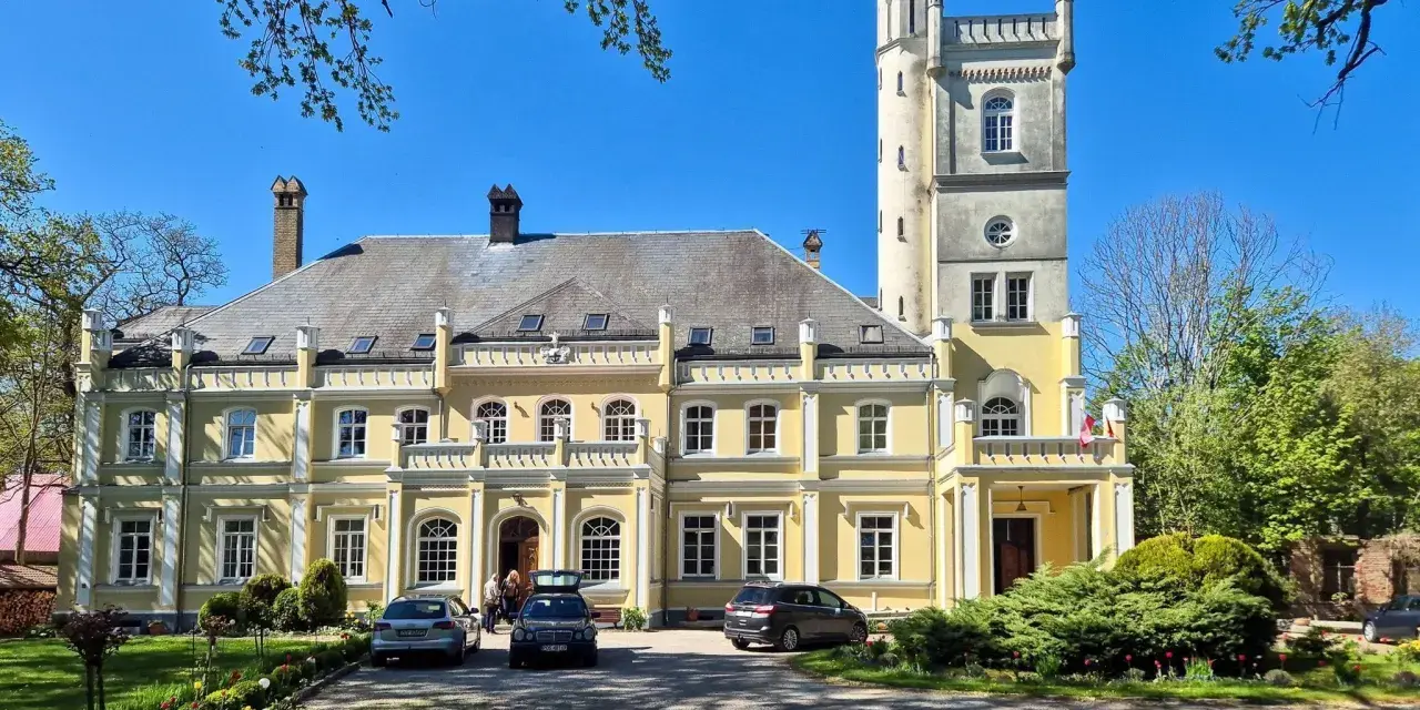 Żółty pałac w Rybokartach z wieżą, otoczony zielenią.