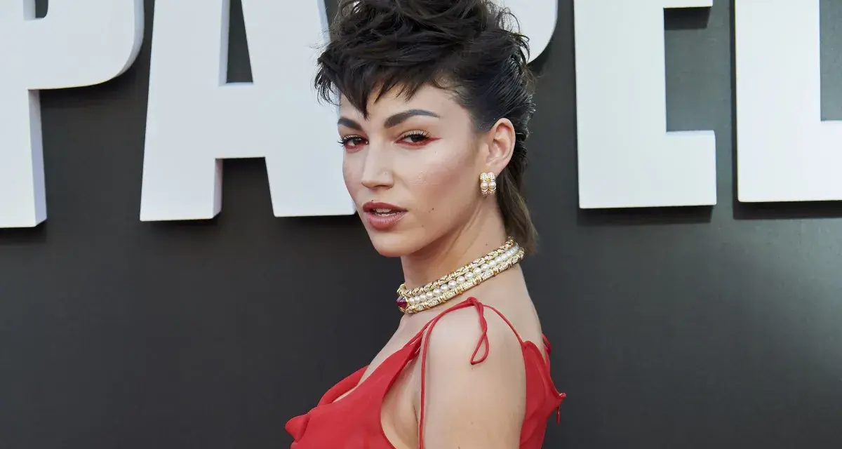 Mujer con un llamativo mullet largo, maquillaje rojo y vestido escarlata, posando ante un fondo con letras blancas.