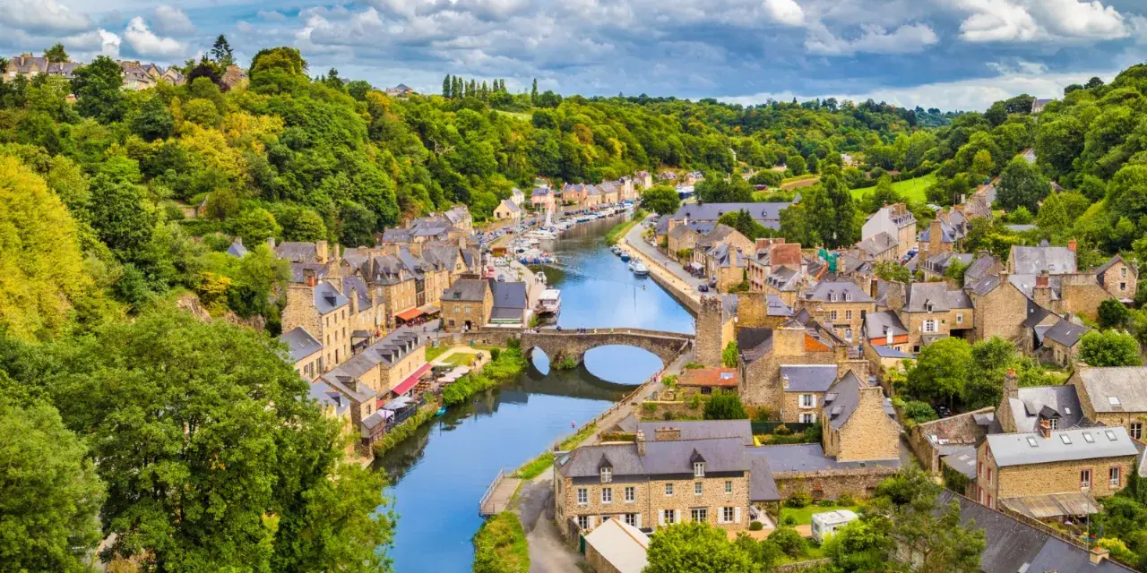 A picturesque view of Dinan, France, with its charming stone buildings lining a winding river, a stone bridge, and lush green hills under a cloudy sky.