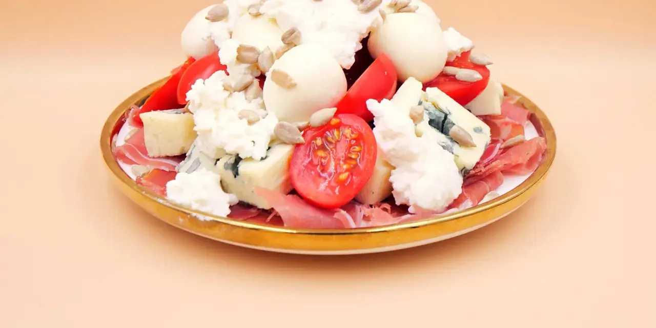 Wykwintna sałatka z szynką parmeńską, serami pleśniowymi, mozzarellą, pomidorkami i słonecznikiem na złotym talerzu.