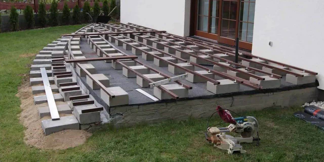 Rozpoczyna się montaż desek tarasowych na legarach. Podstawa z bloczków betonowych i podsypki piaskowej czeka na wykończenie.