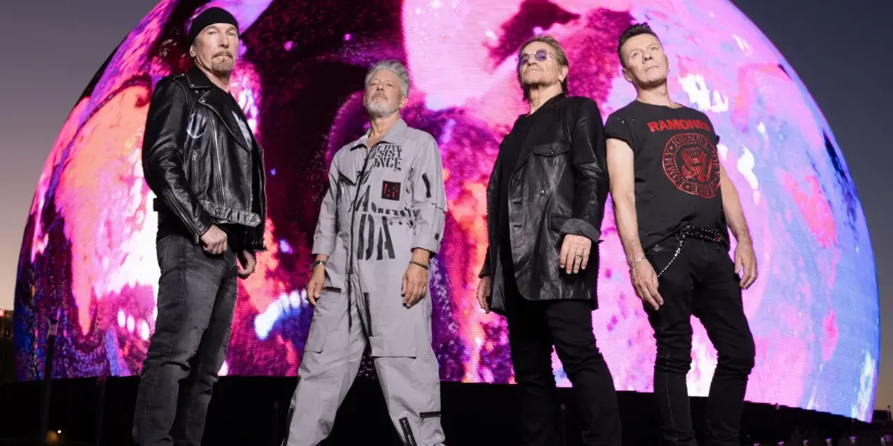 U2 w nowej płycie. Czterech członków zespołu stoi przed gigantycznym, kolorowym ekranem.
