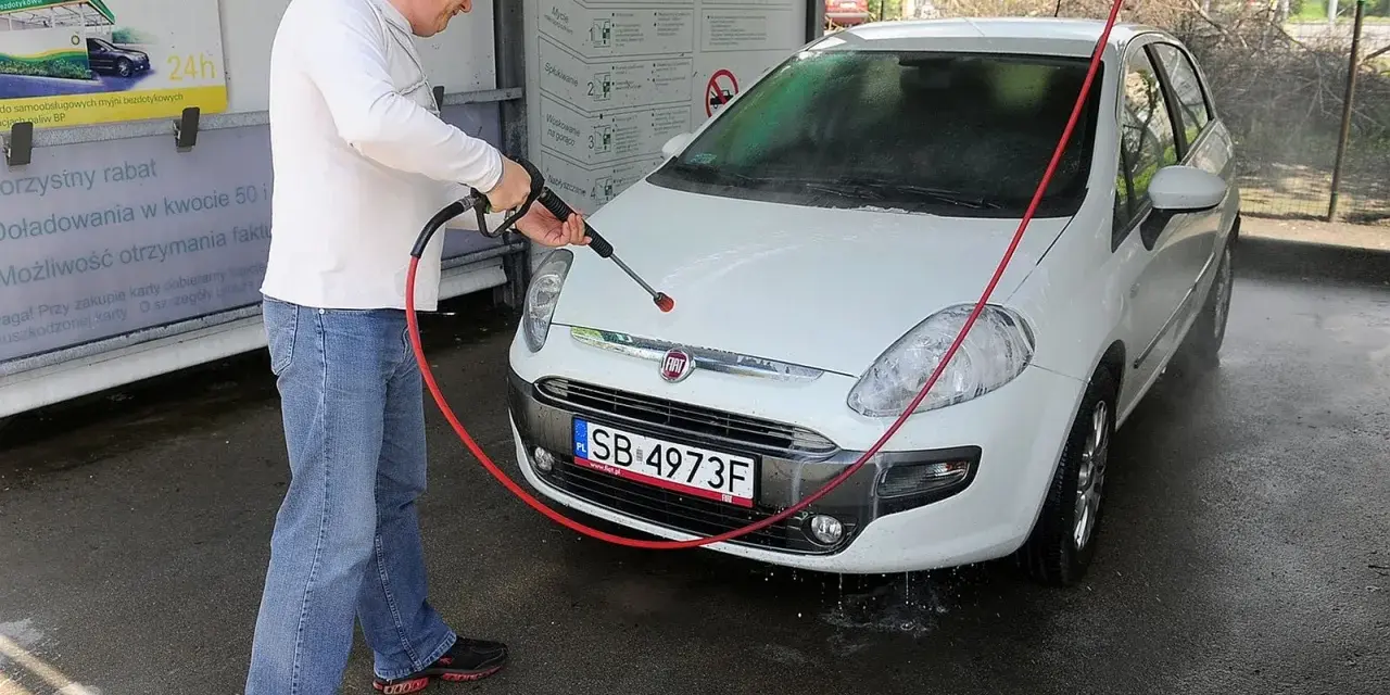 Mężczyzna myje białe auto na myjni. Na tablicy informacyjnej widnieje napis "Możliwość otrzymania faktury", sugerujący, że na myjni można płacić kartą.