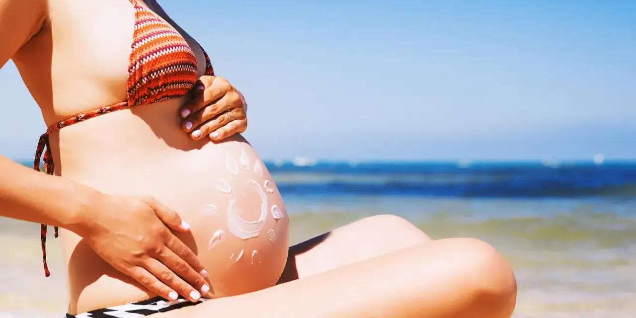 Ciężarna kobieta na plaży, z kremem przeciwsłonecznym na brzuchu w kształcie słońca. Bezpieczne opalanie natryskowe w ciąży.