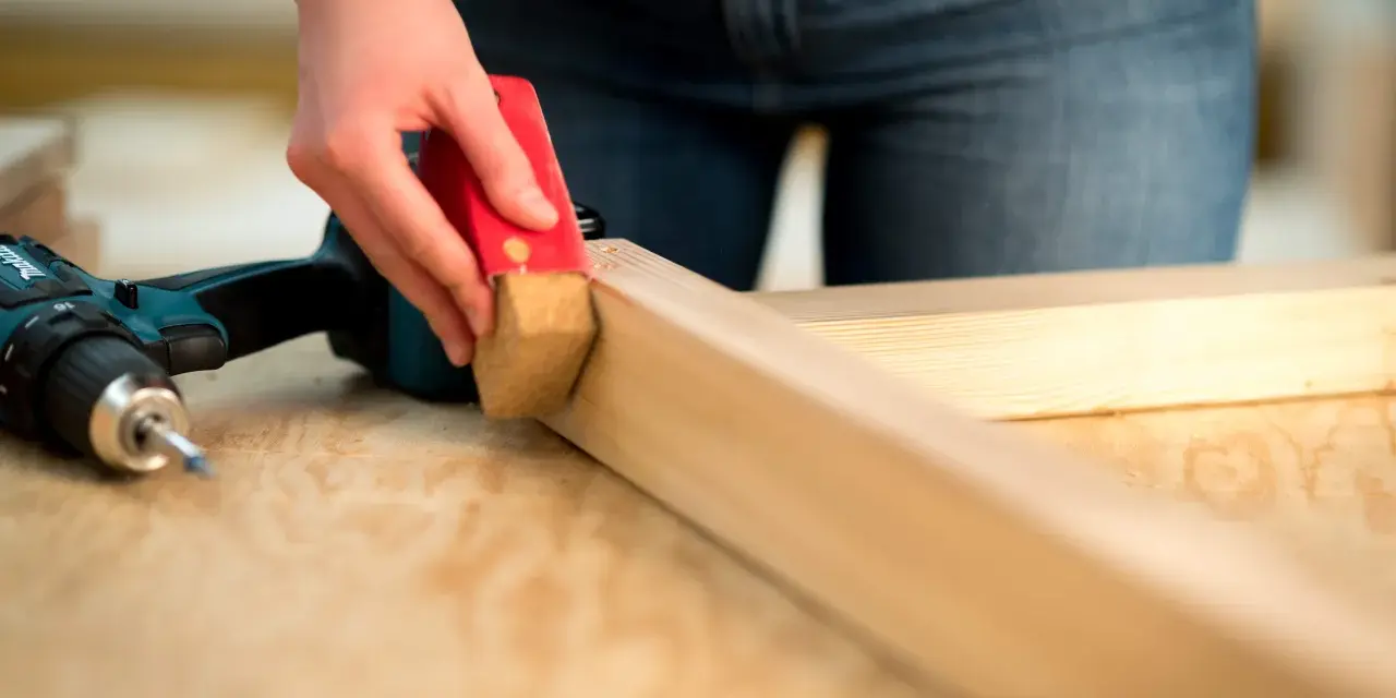 Eine Hand schleift mit einem roten Schleifklotz ein Holzstück. Daneben liegt ein Akkuschrauber.