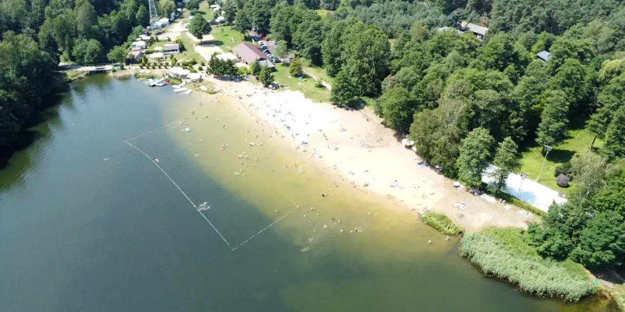 Słoneczny dzień nad jeziorem Rogoźnik. Ludzie kąpią się i opalają na piaszczystej plaży, otoczonej zielonym lasem.