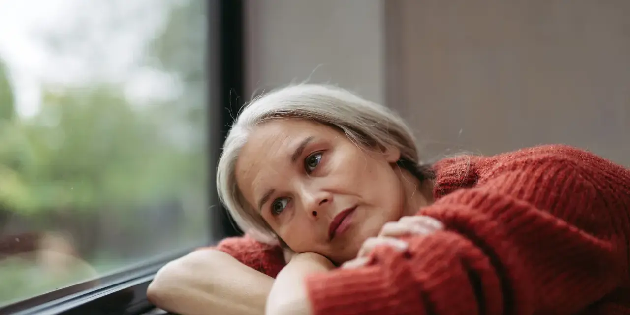Donna con capelli grigi guarda fuori dalla finestra, pensierosa. Potrebbe cercare rimedi per la depressione estiva.