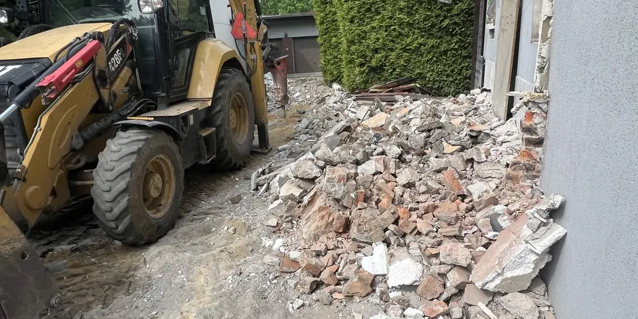 Żółta koparko-ładowarka pracuje przy wyburzeniu domu. Gruz i cegły leżą na ziemi. Koszt wyburzenia domu jest wysoki.
