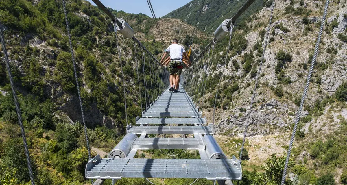Un ponte tibetano in Puglia attraversa una gola verdeggiante, con persone che camminano su passerelle metalliche sospese tra montagne.
