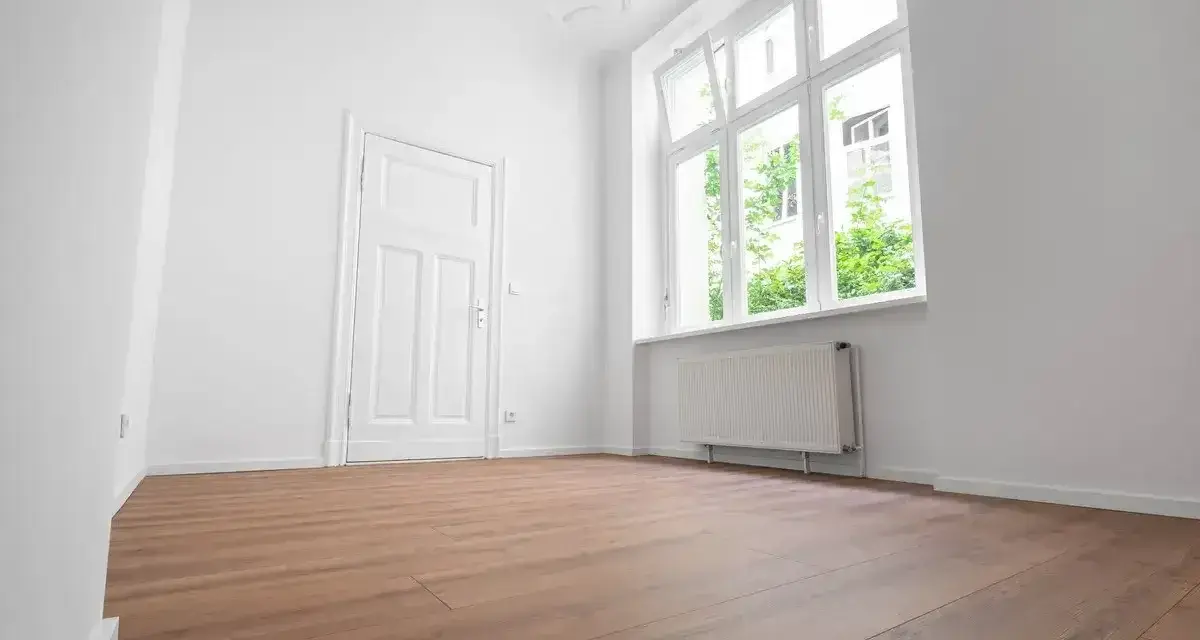 Puste, jasne pomieszczenie z białymi ścianami, drzwiami i oknem. Drewniana podłoga i grzejnik sugerują, że to gotowe do zamieszkania mieszkanie socjalne.