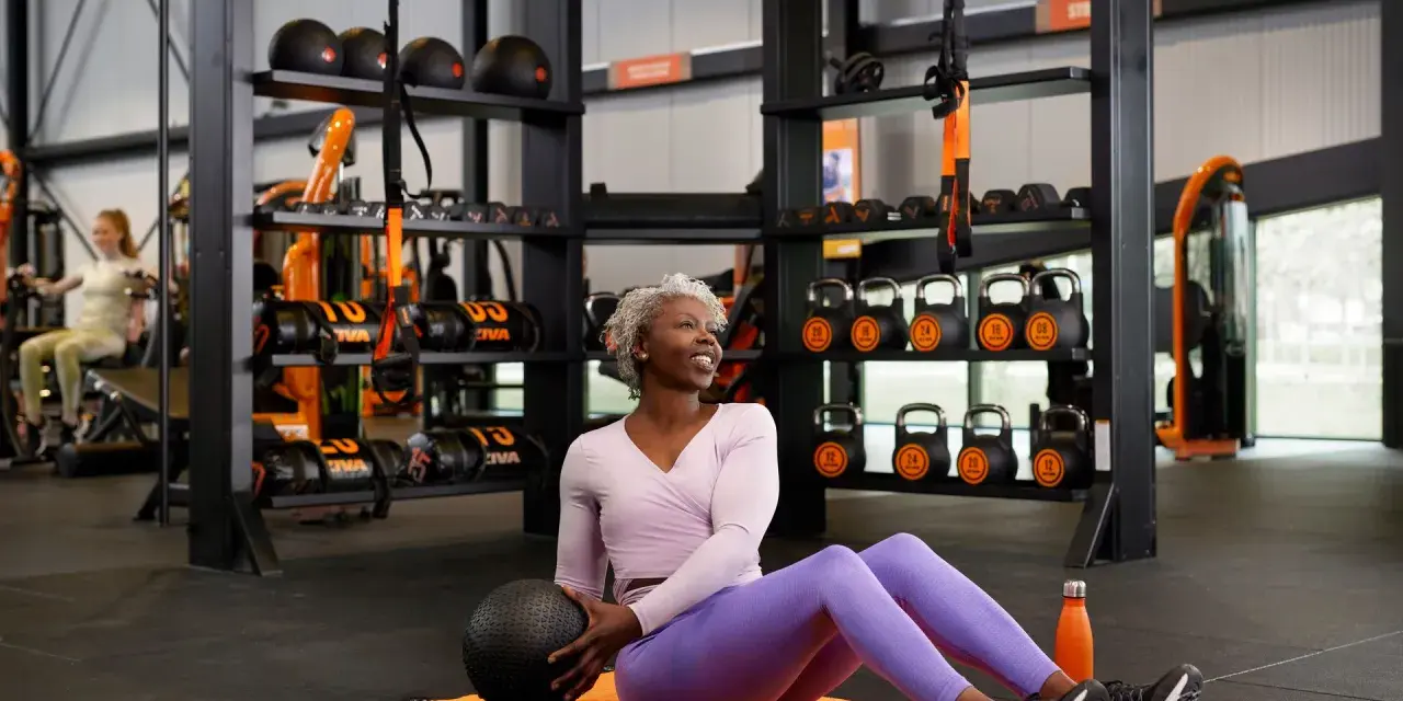 Une femme souriante fait un réglage photo sport en salle, tenant un ballon de médecine. L'équipement de gym est visible en arrière-plan.
