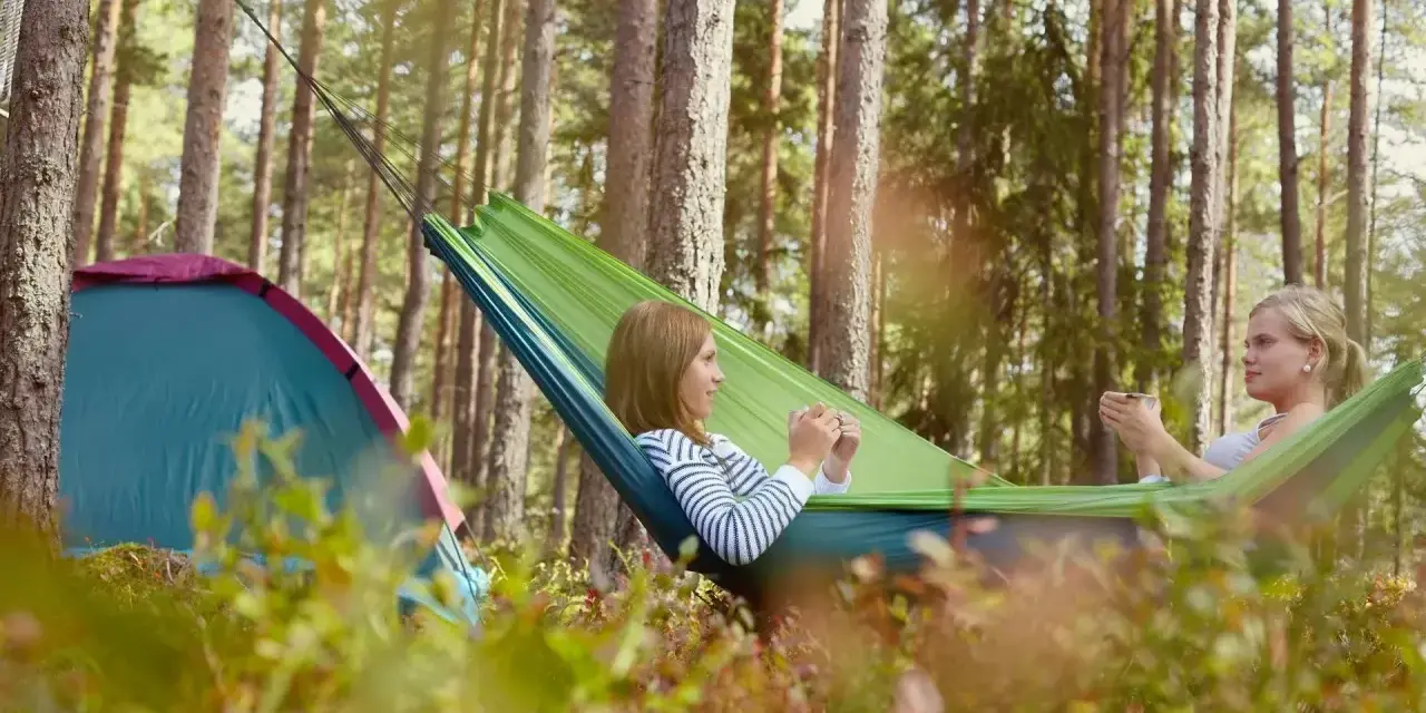 Två tjejer njuter av en lugn stund i en hängmatta på en av Sveriges fina campingar. Bredvid dem står ett tält.