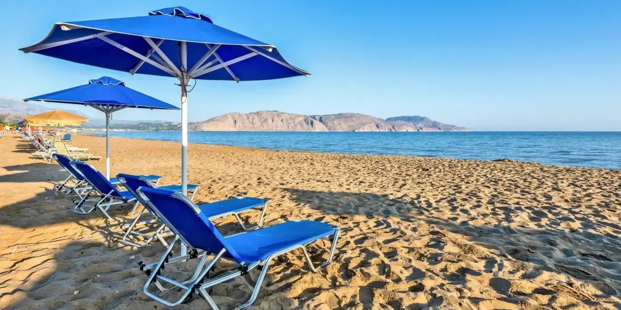 Błękitne leżaki i parasole na piaszczystej plaży Kavros Beach, z widokiem na spokojne morze i górzyste wybrzeże w oddali.