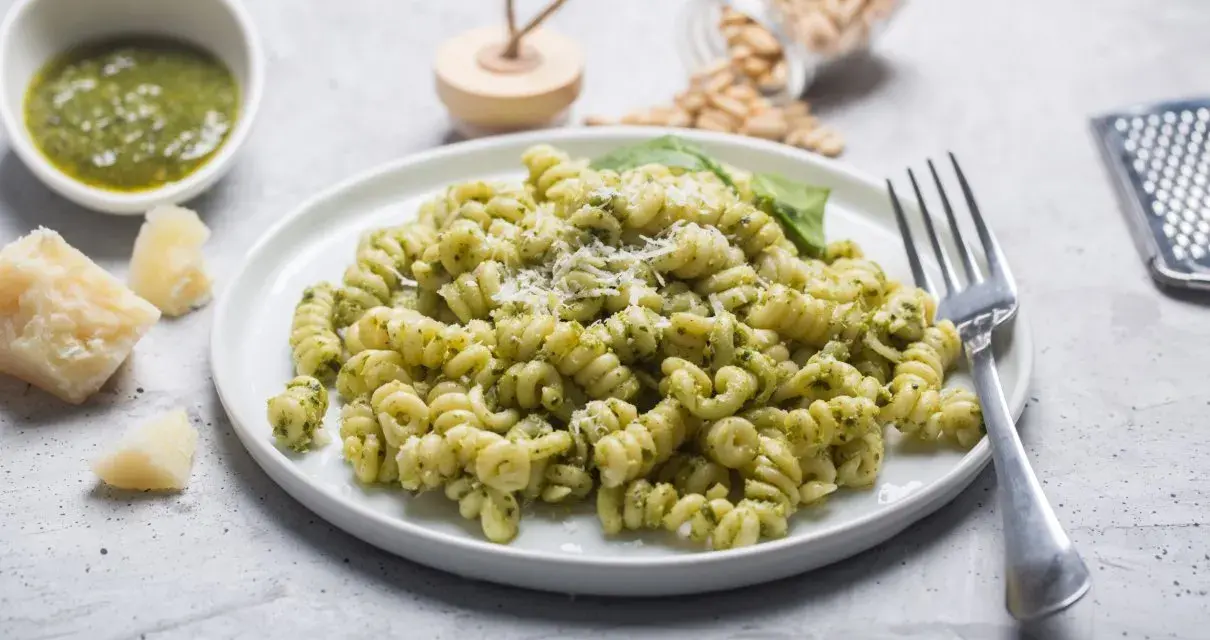 Pyszny makaron z pesto ze słoika i śmietaną, posypany parmezanem. Obok miseczka z pesto i kawałki sera.
