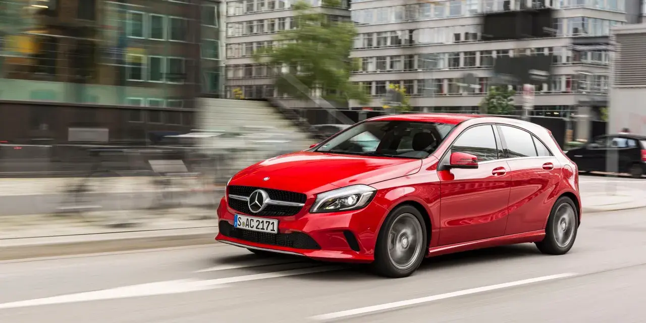 Czerwony Mercedes-Benz W176 pędzi ulicą miasta, mijając nowoczesne budynki.
