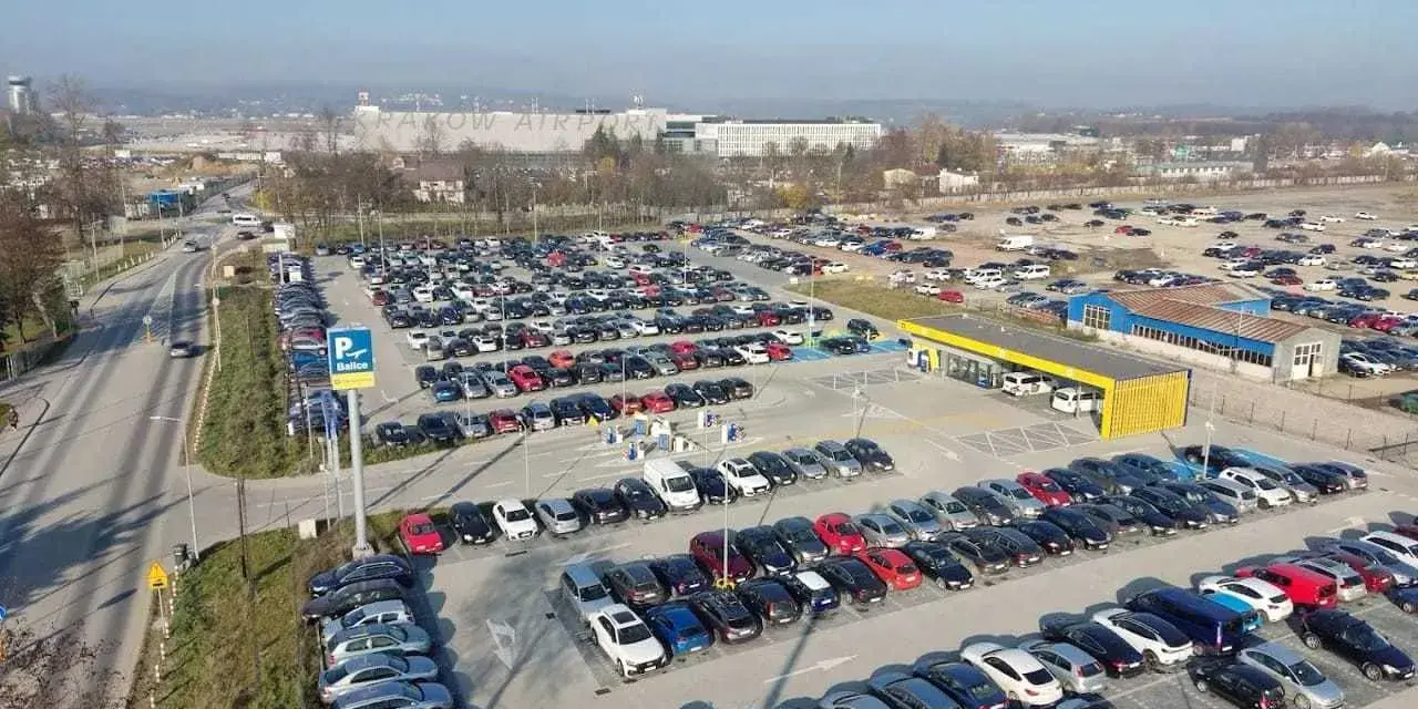 Duży parking przy krakow katowice airport. Wiele samochodów zaparkowanych w rzędach, w tle budynki lotniska.