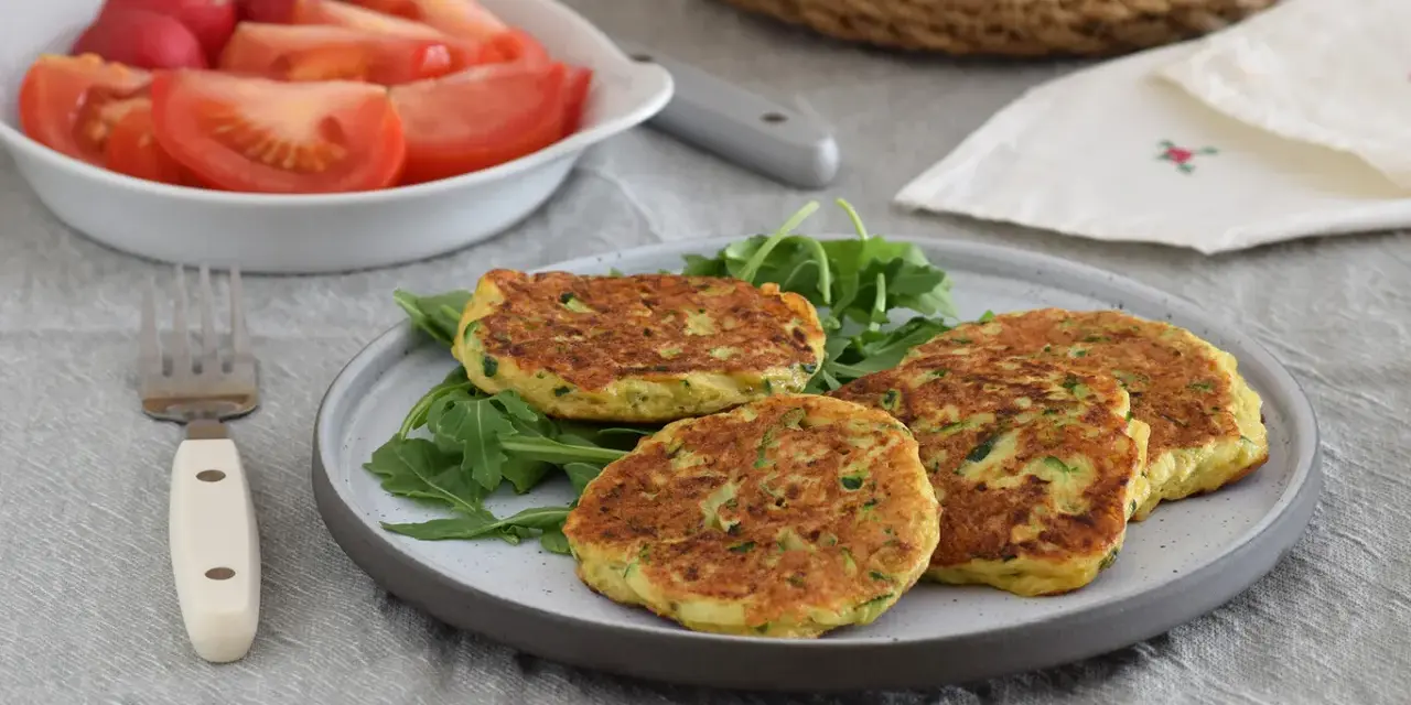 Tortitas de calabacín doradas y esponjosas, servidas sobre rúcula fresca. Acompañadas de una ensalada de tomate y rábano.