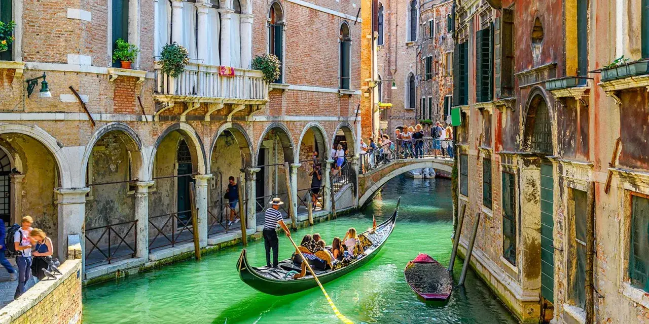 Un gondoliere a Venezia guida una gondola con turisti lungo un canale verde, tra palazzi storici e un ponte affollato.