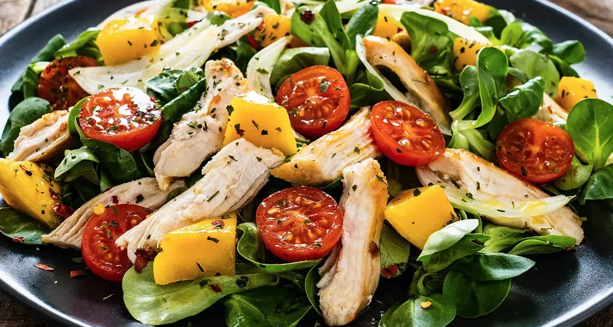 Pyszna sałatka z kurczaka z pomidorkami koktajlowymi i kawałkami mango, podana na ciemnym talerzu.