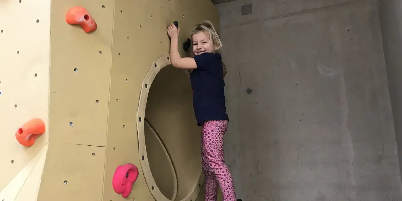 Ein fröhliches Mädchen klettert in einer Kletterhalle für Kinder. Sie lächelt und hält sich an einem Griff fest.