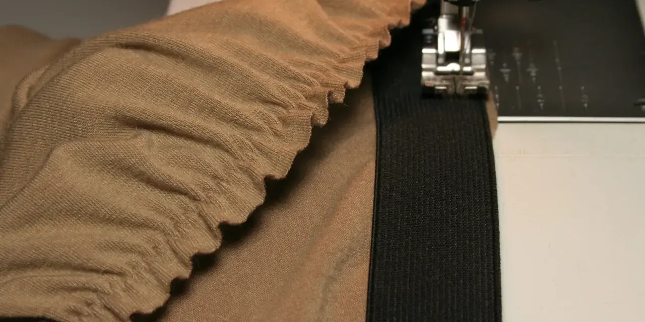 Machine à coudre en train de coudre un élastique sur un tissu beige froncé. Le pied presseur est visible, ainsi que l'aiguille.