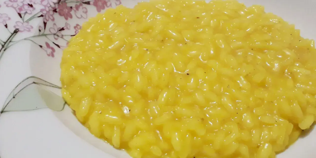 Un cremoso risotto allo zafferano, servito in un piatto bianco decorato con fiori rosa e rami verdi.