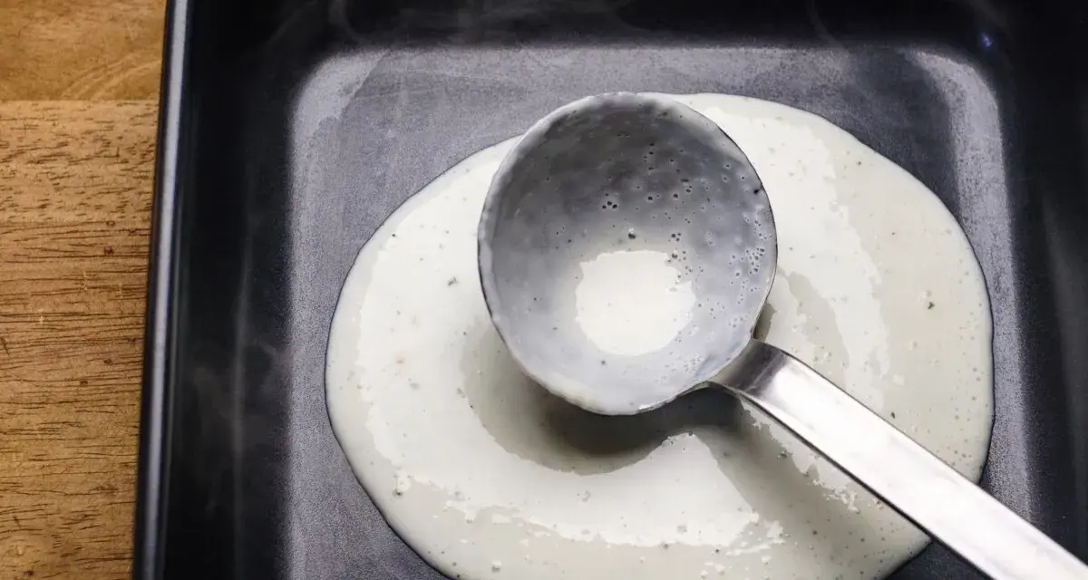 Eine Schöpfkelle verteilt cremige Bechamel-Sauce in einer schwarzen Auflaufform.