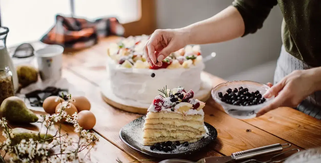 Manos expertas decoran un pastel en una mesa de reposteria y pasteleria, con frutas frescas y arándanos.