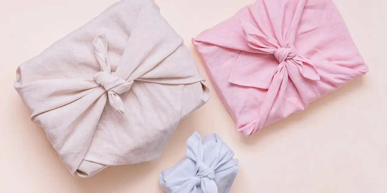 Trois paquets cadeaux joliment emballés dans des tissus de différentes couleurs (beige, rose, bleu clair). Lequel tissu pour furoshiki ? Le lin semble idéal.