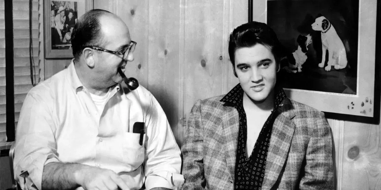 Elvis Presley und Colonel Parker, sein Manager, sitzen nebeneinander. Parker raucht Pfeife, Elvis blickt in die Kamera.