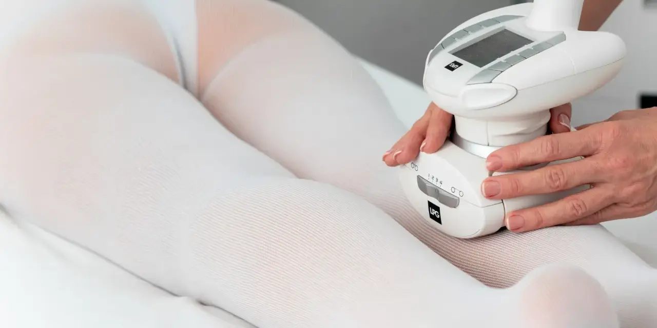 Zabieg endermologii w domu. Specjalistka wykonuje masaż specjalistyczną głowicą na nogach pacjentki w białej bieliźnie.