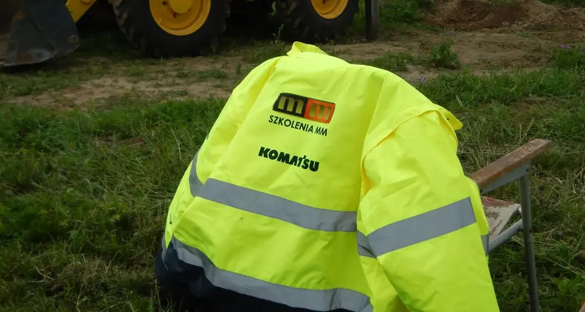 Żółta kurtka z logo "SZKOLENIA MM KOMATSU" leży na krześle. W tle koparko-ładowarka. Idealna na pytania na koparko ładowarkę.