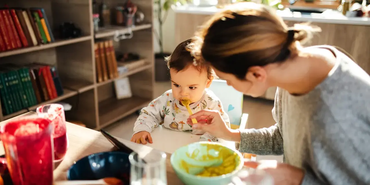 Mama karmi łyżeczką 6 miesięczne dziecko. Na stole są owoce i napoje.