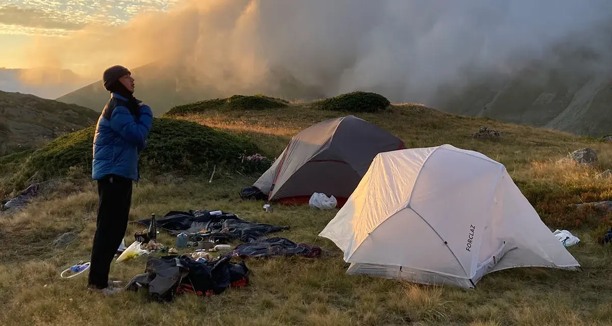 Une personne se tient près de tentes dans une aire de bivouac, profitant du lever du soleil sur les montagnes.