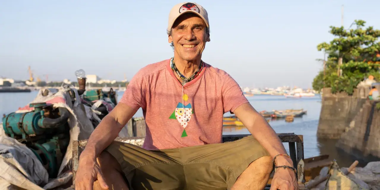 Le chanteur Mano Negra, souriant, assis sur un banc rouillé, avec un collier coloré et un t-shirt à motif poisson.