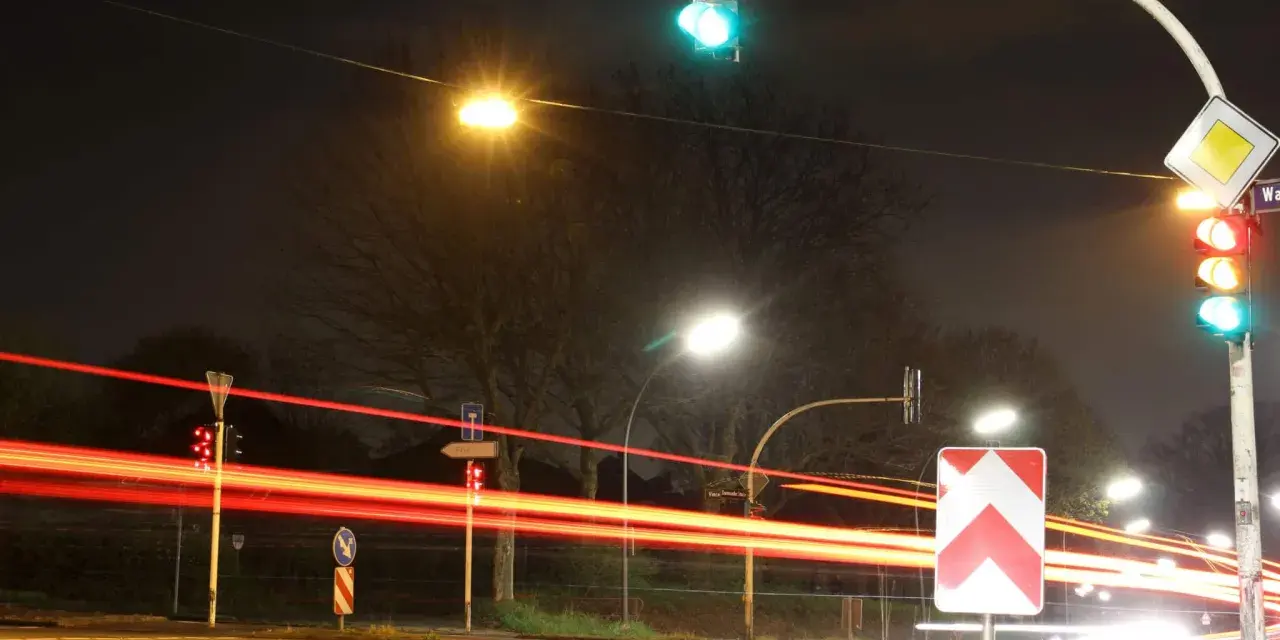 Nachts an einer Kreuzung mit roten Lichtspuren von Autos. Ampeln zeigen Grün und Rot. Ein Vorfahrtsschild und ein Richtungspfeil sind zu sehen. 132 bkat.