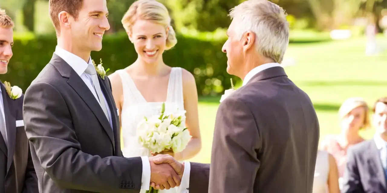 Pan młody ściska dłoń ojca panny młodej, otrzymując jego blogoslawienstwo rodzicow na slub. Panna młoda uśmiecha się z bukietem.