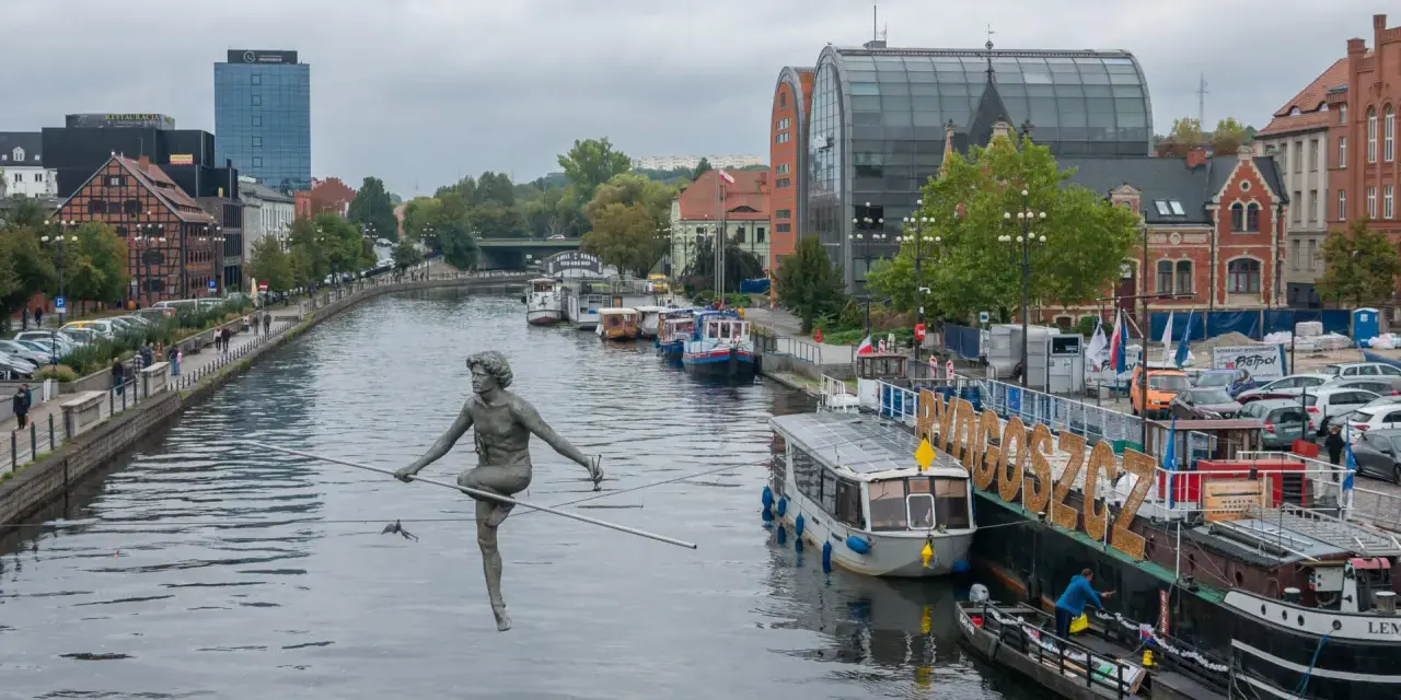 Bydgoszcz atrakcje: rzeźba na linie nad Brdą, cumujące statki i budynki nad rzeką.