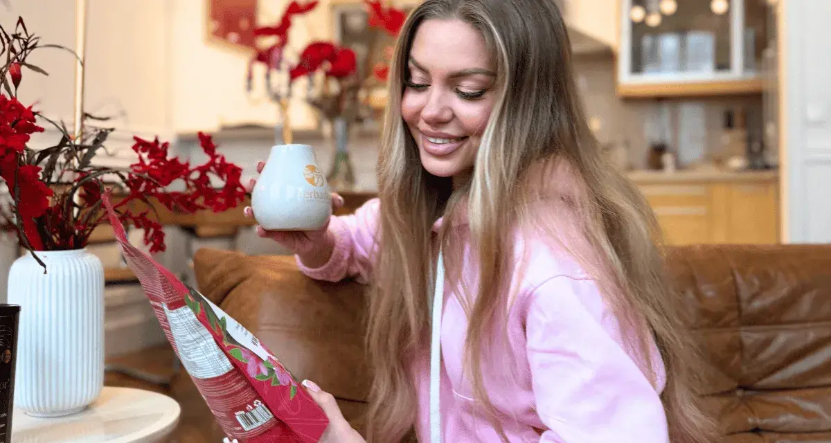 Uśmiechnięta kobieta trzyma kubek i paczkę yerba mate, przygotowując się do parzenia.