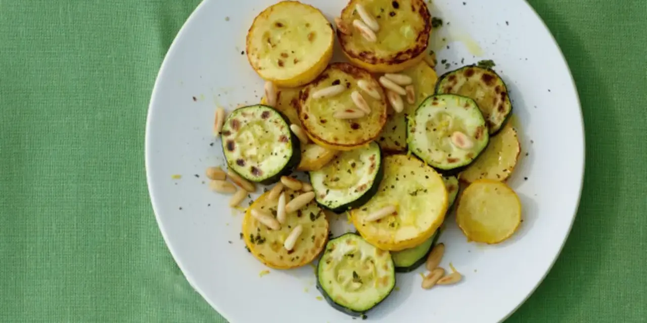 Gegrillte gelbe Zucchini-Scheiben mit Pinienkernen und Kräutern auf einem weißen Teller.