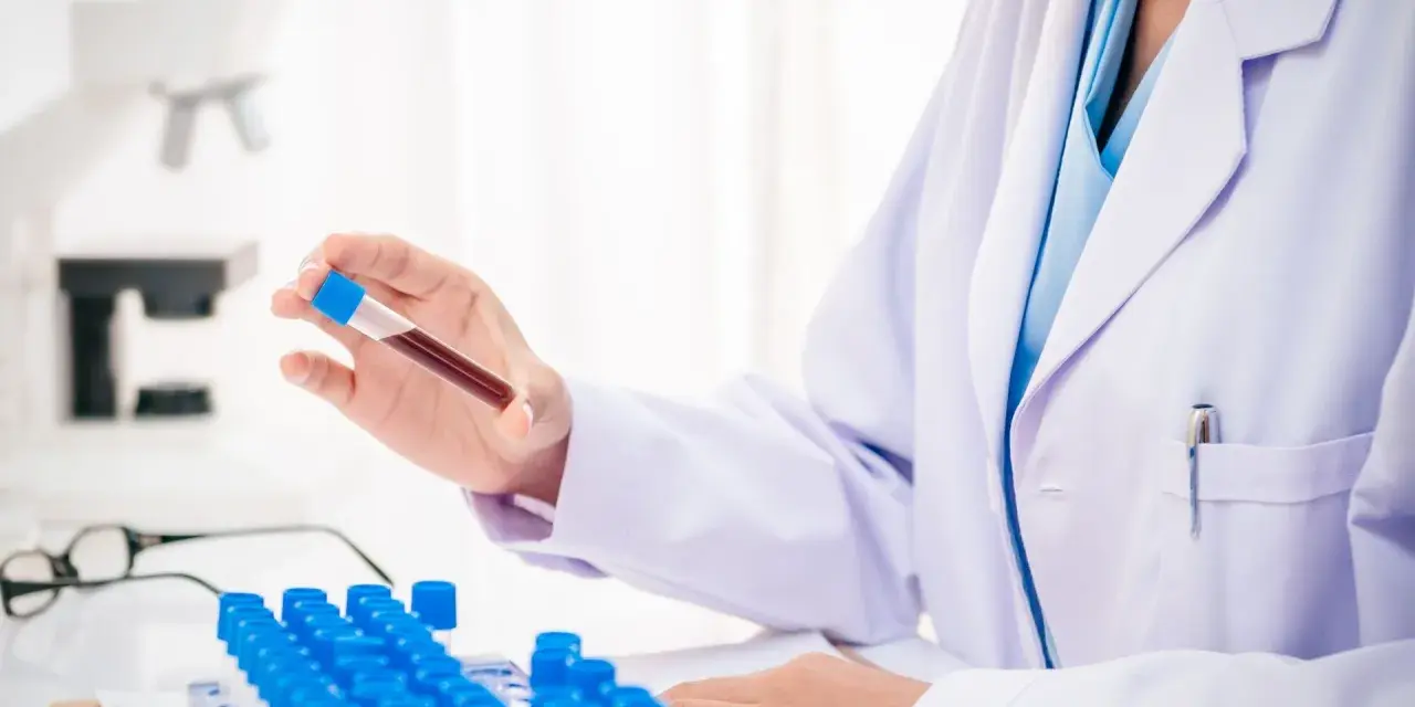 Laborant w białym fartuchu trzyma probówkę z krwią, w tle mikroskop i rząd probówek.
