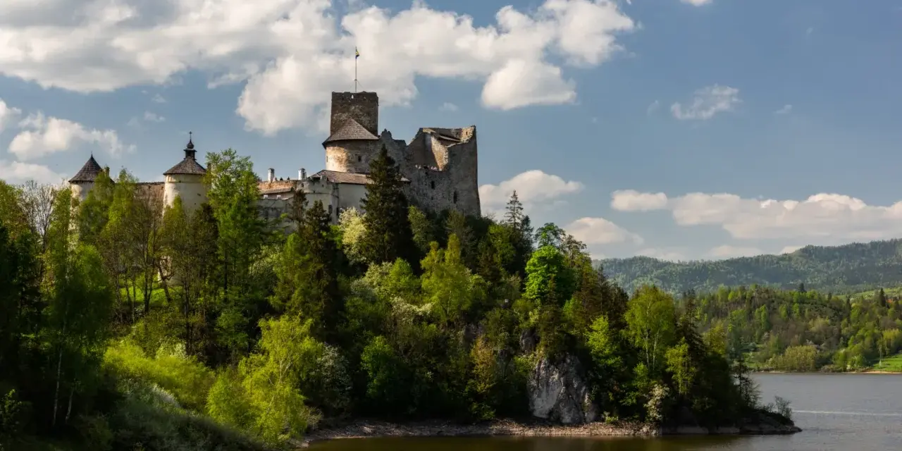 Zamek w Czorsztynie góruje nad jeziorem, otoczony zielenią. W oddali widać góry.