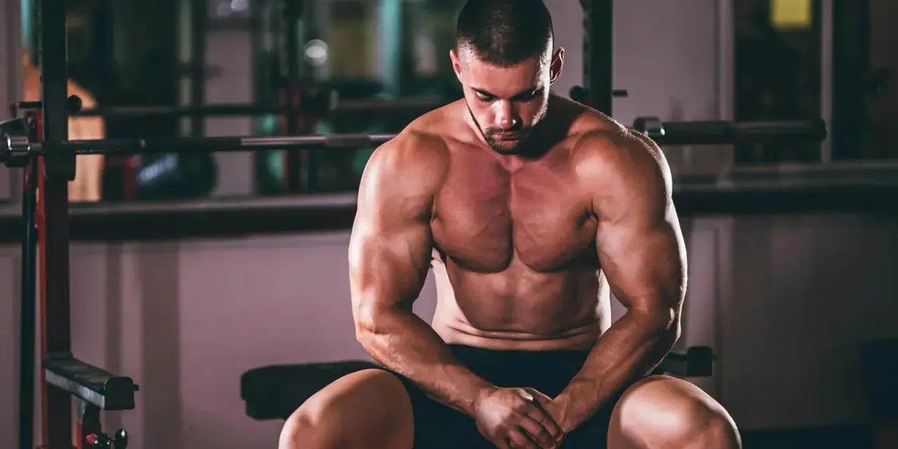 Un homme musclé, concentré, s'entraîne pour prendre du muscle rapidement. Il est assis dans une salle de sport, le regard baissé.