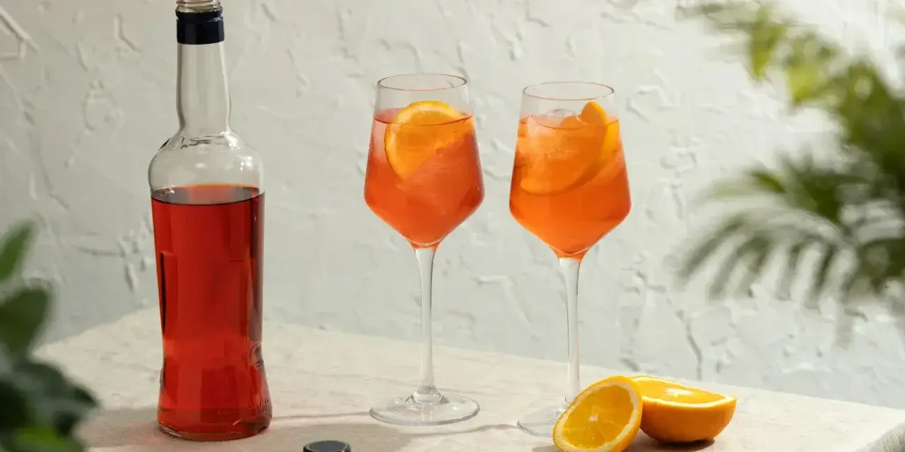 Idealny aperol spritz przepis: butelka Aperolu, dwa drinki z pomarańczą i lodem, obok połówki pomarańczy.