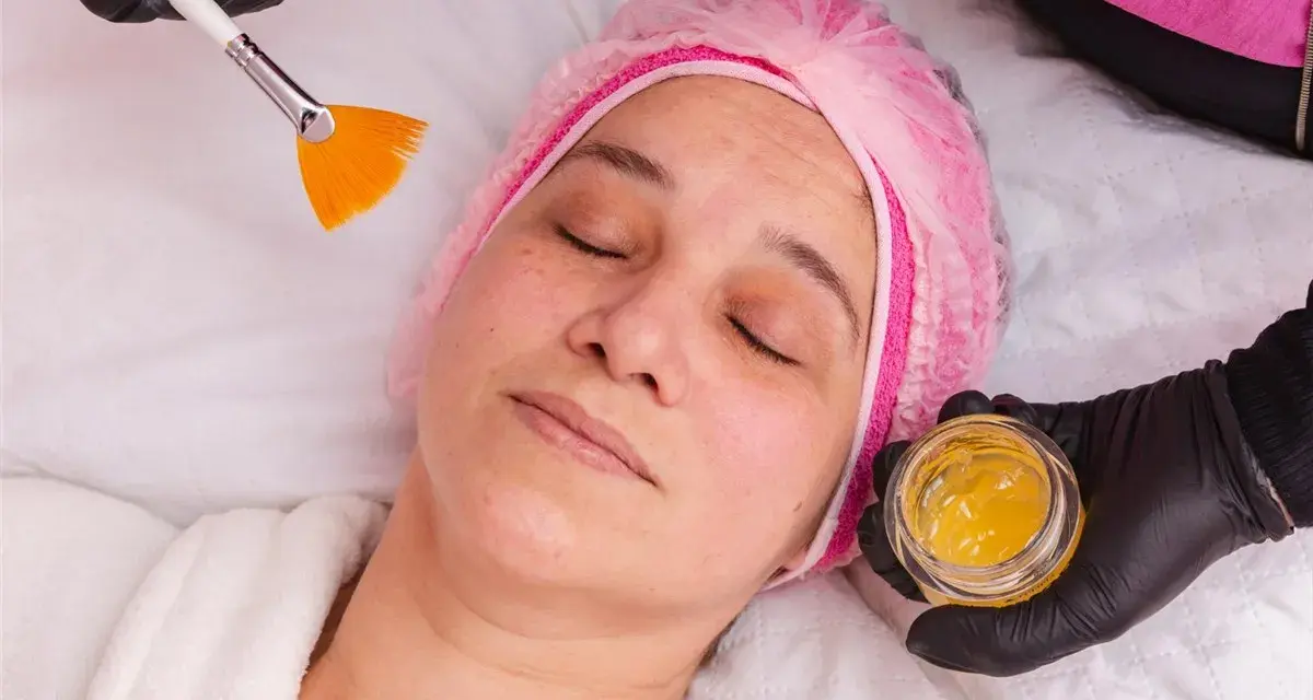 Kobieta w czepku leży z zamkniętymi oczami, a kosmetyczka przygotowuje maseczkę z miodu na twarz, używając pędzelka.