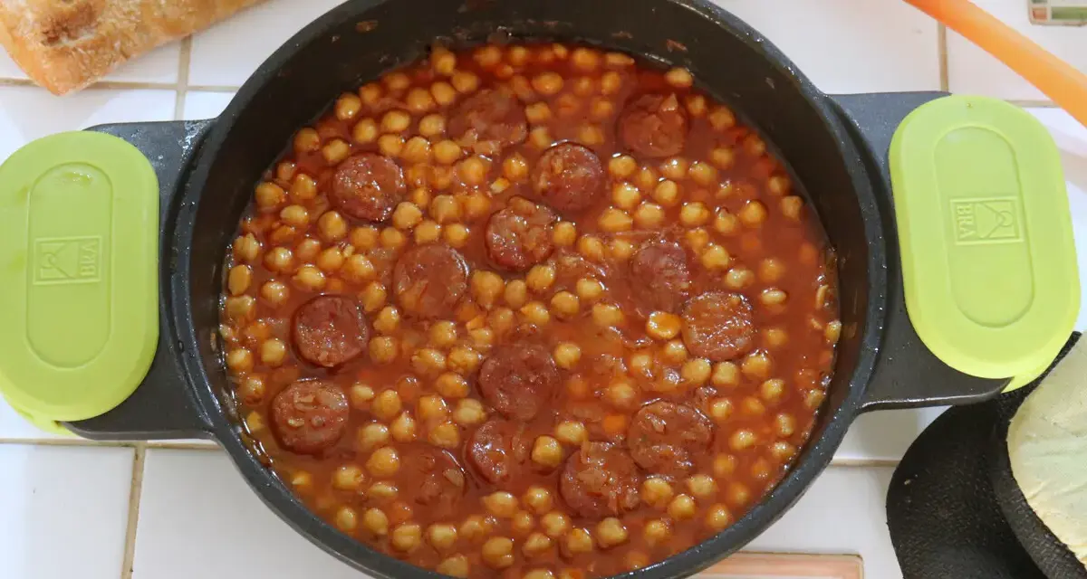 Guiso de garbanzos con chorizo en una cazuela, con pan y cuchara naranja.