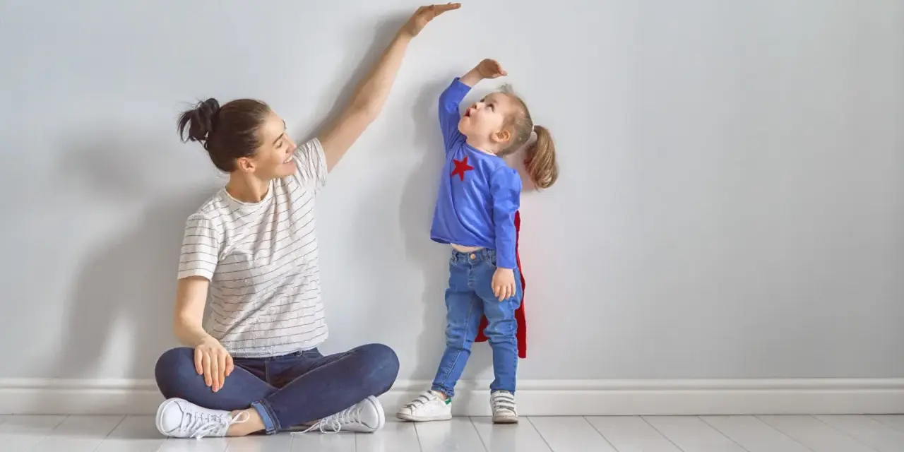 Mama mierzy wzrost córeczki w stroju superbohaterki. To właśnie wtedy dzieci rosną najszybciej, zaskakując nas każdego dnia.